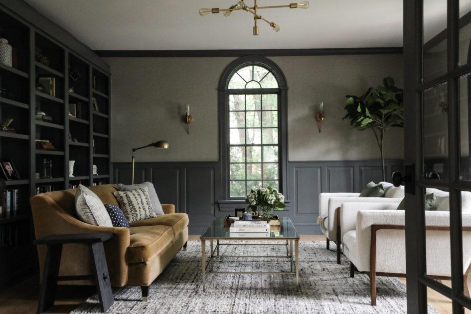 A sophisticated living room with dark gray walls featuring a large arched window. It is furnished with a brown sofa, two white armchairs, a glass coffee table, and shelves filled with books and decor. A potted plant and modern chandelier add elegance to the space.