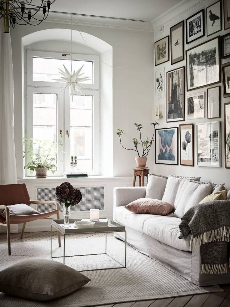 A cozy living room with a large window, white sofa, and a variety of framed pictures on the wall. A coffee table with flowers and a candle sits in front of the sofa. There's a wooden chair with a pink cushion, a soft rug, and plants adding a touch of greenery.