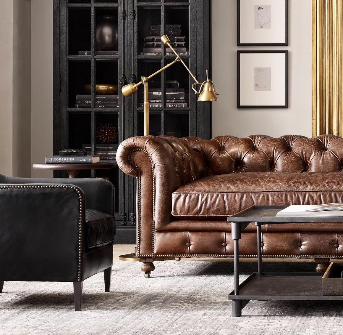 A cozy living room with a tufted brown leather sofa and a black leather armchair, both accented with brass nailhead trim. In the background, there's a black wooden cabinet with glass doors, and a brass floor lamp with two adjustable lights. Framed artwork hangs on the wall.