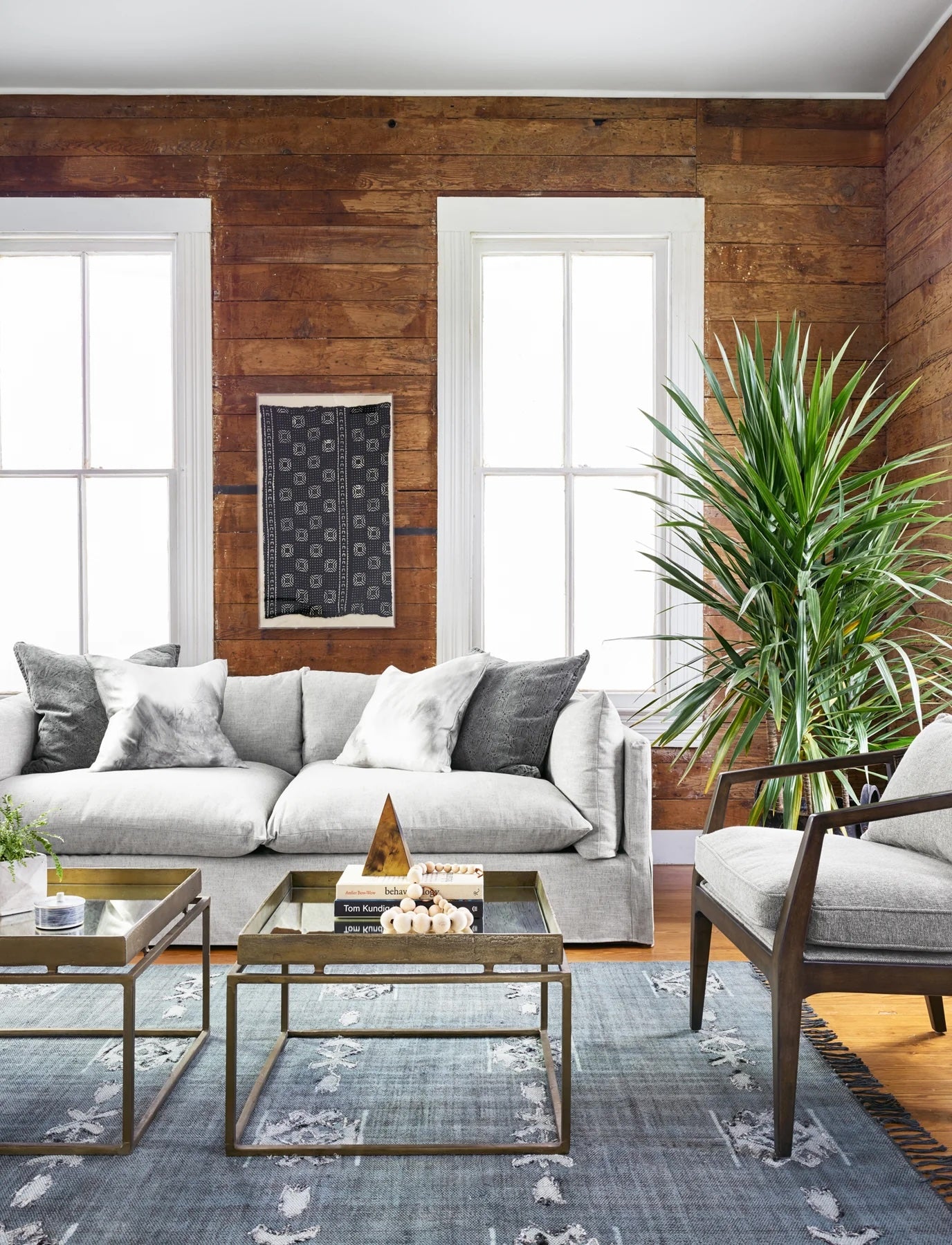 A modern living room with rustic wooden walls features a light grey sofa and a matching armchair. A large green plant stands next to the armchair. A dark grey area rug, two coffee tables with books and decorations, and bright natural light complete the look.