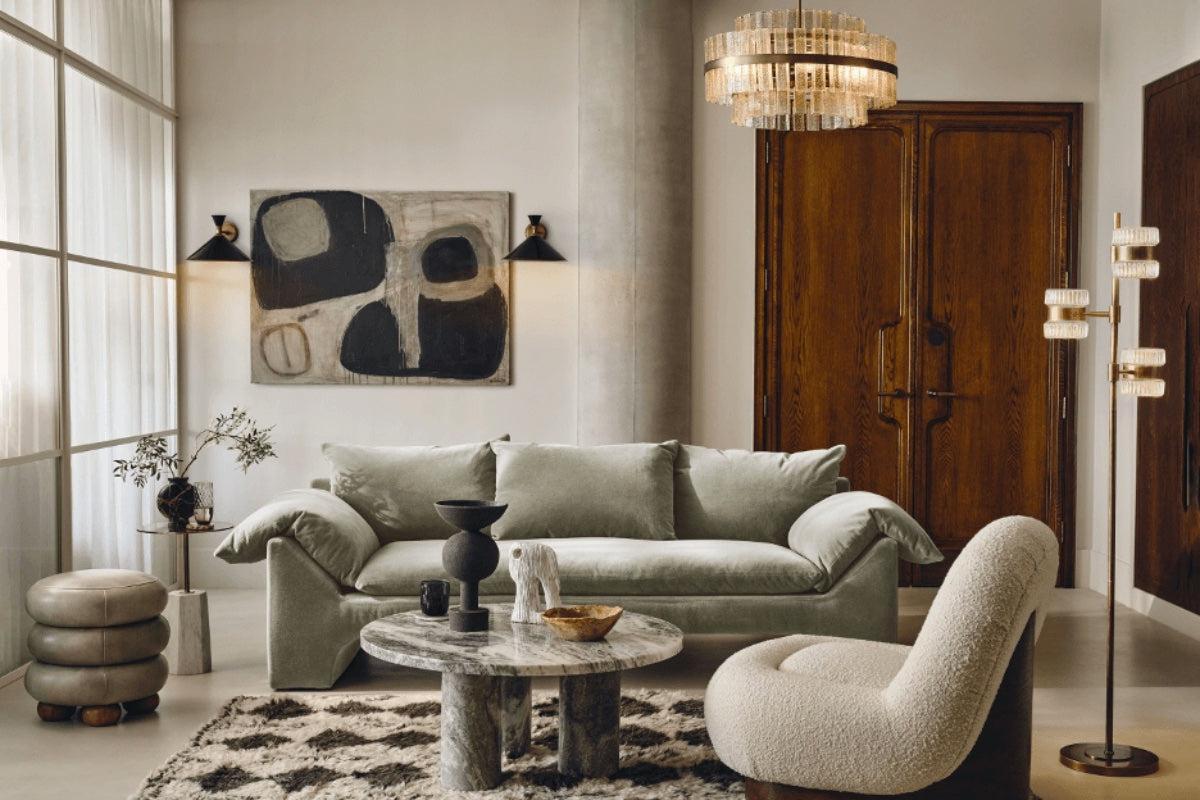 A modern living room with a plush, light gray sofa, a round marble coffee table, and a cream-colored lounge chair on a geometric-patterned rug. A large abstract painting, elegant floor lamp, decorative items, and wooden double doors adorn the space.