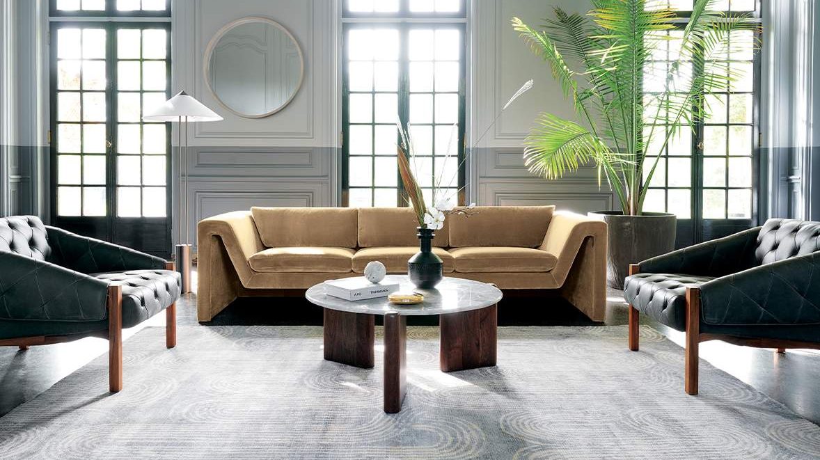 A modern living room featuring a mustard yellow sofa against a grey paneled wall with tall windows. Two black leather chairs face a round marble coffee table in the center, which is adorned with decorative items and a vase. A large potted plant adds greenery.