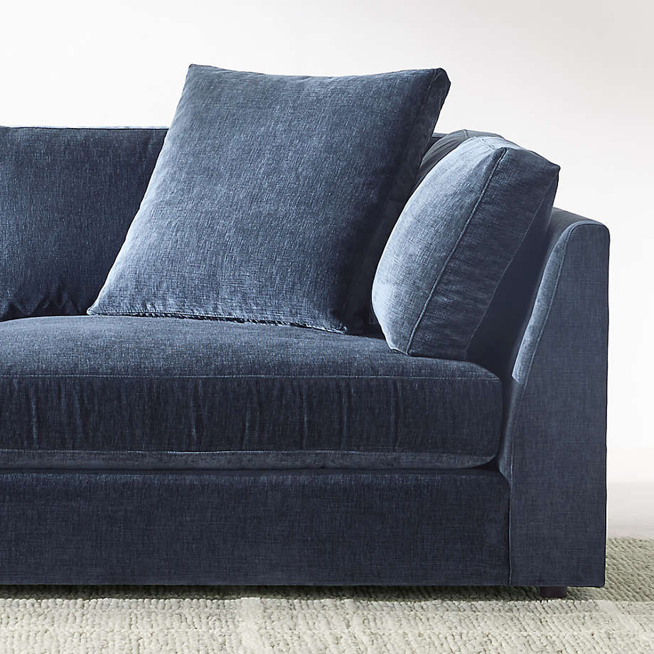 A Monterey Modular 3 Piece L Shaped Sectional Sofa, crafted in plush dark blue velvet and accented with two large square cushions, is positioned against a light gray wall. The textured fabric enhances the depth of the scene, and the sofa rests on a light beige carpet, creating a cozy and modern living space.