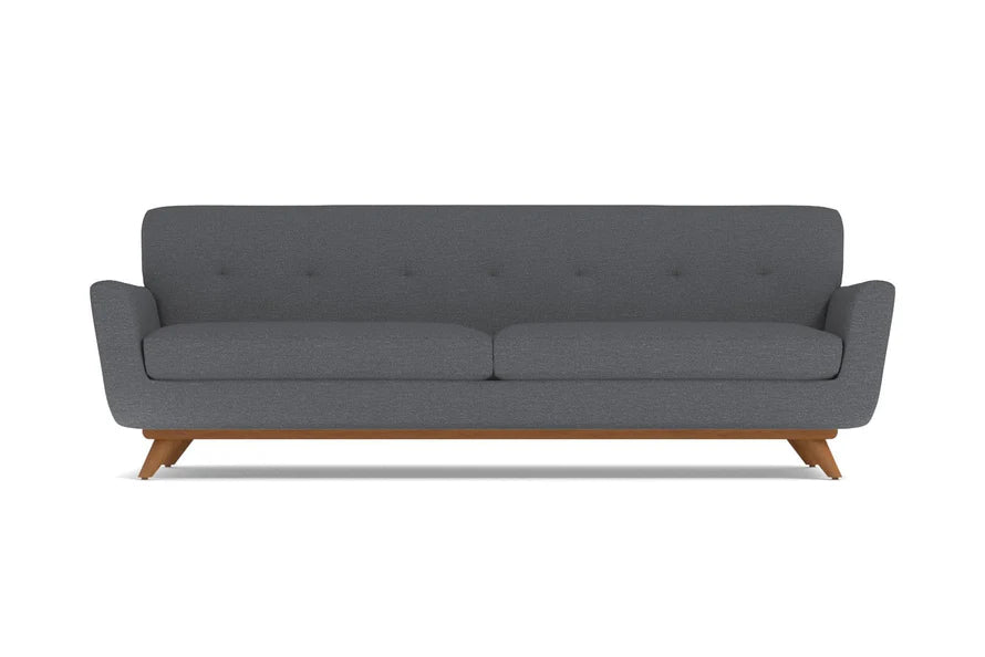 The Carson Sofa offers a modern aesthetic with its dark grey hue and minimalist design. It features a cushioned back adorned with subtle button detailing, sleek armrests, and is supported by mid-century styled wooden legs.