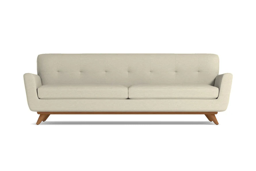 The Carson Sofa is a mid-century modern design featuring a beige color with a tufted backrest and two seat cushions. It is supported by a wooden base and angled legs, all presented against a plain white background.
