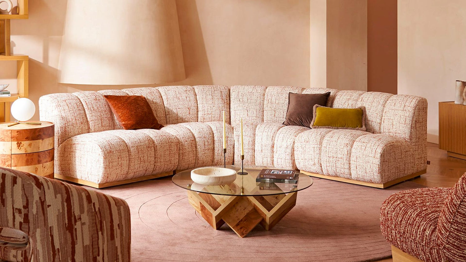A warmly lit living room featuring a peach-colored, textured sectional sofa with various pillows, including a brown and a mustard yellow one. A round glass coffee table with a geometric wooden base sits on a circular area rug. Shelves and a lamp are in the background.