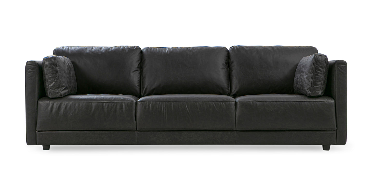 The Domus 93" Leather Sofa in chestnut is displayed, featuring a modern design with straight lines, three seat cushions, and two matching throw pillows. It incorporates a sleek and minimalistic style with a low backrest.