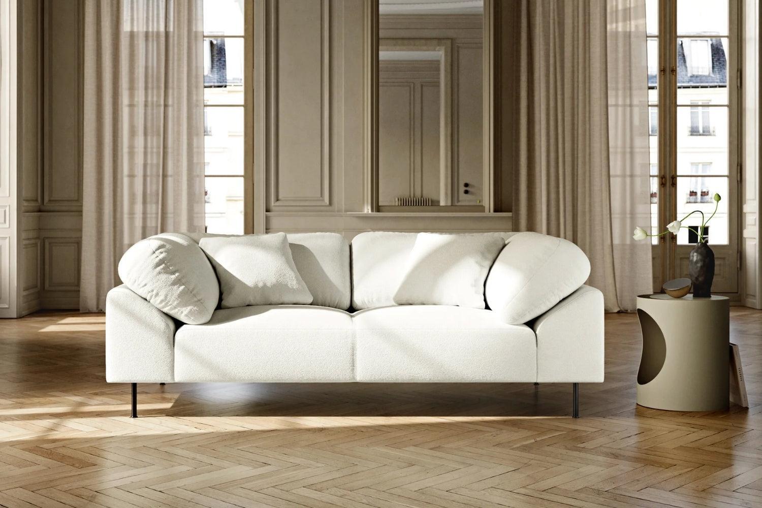 A modern living room features a large, plush white sofa with matching cushions on a wooden herringbone floor. Behind it are floor-to-ceiling windows with sheer curtains. A round side table with a decorative vase stands to the right.