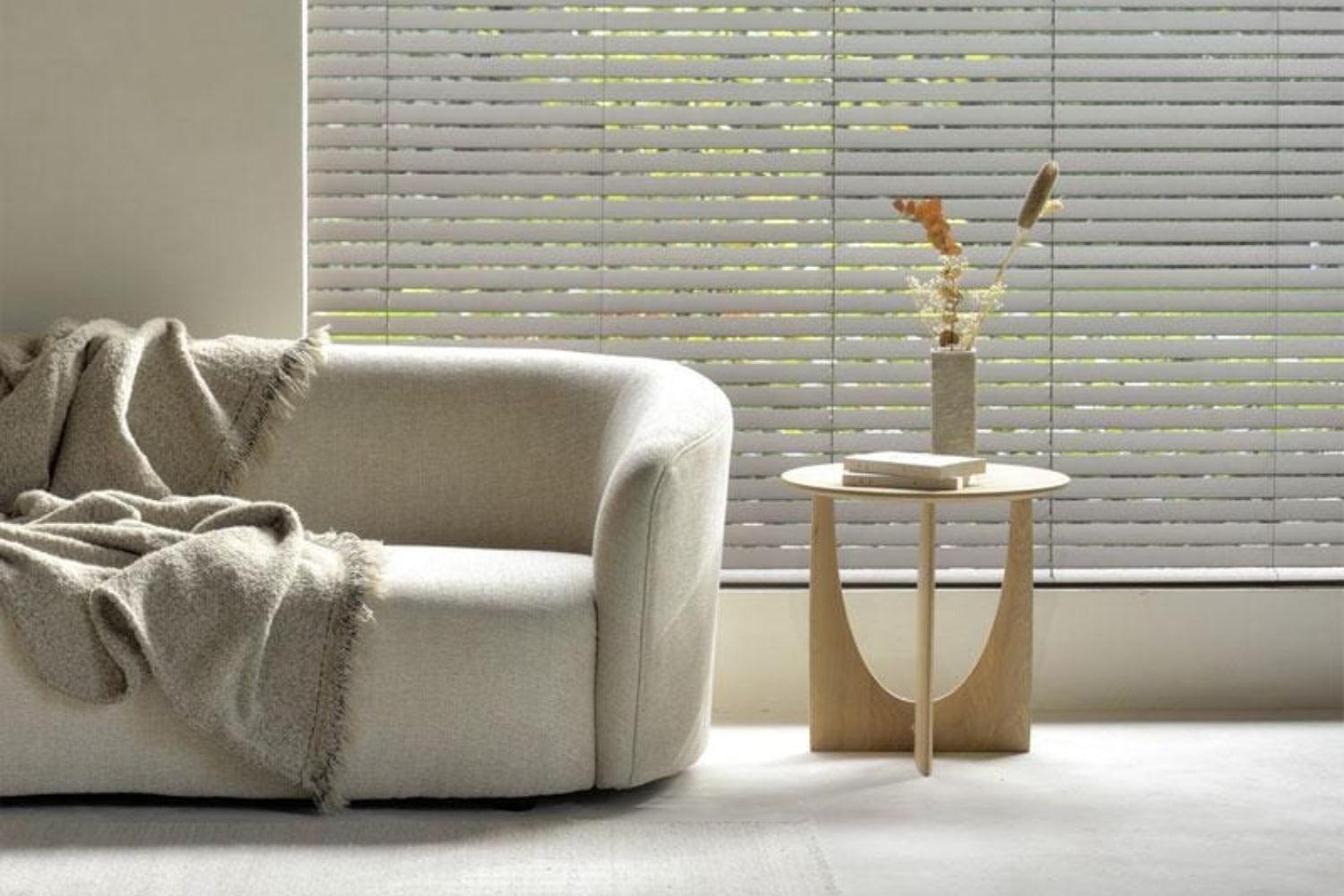 A cozy living room features a plush beige sofa draped with a soft, fringed blanket. Beside it, a minimalist wooden side table holds a dried floral arrangement. Large windows with horizontal blinds let in natural light, creating a warm atmosphere.