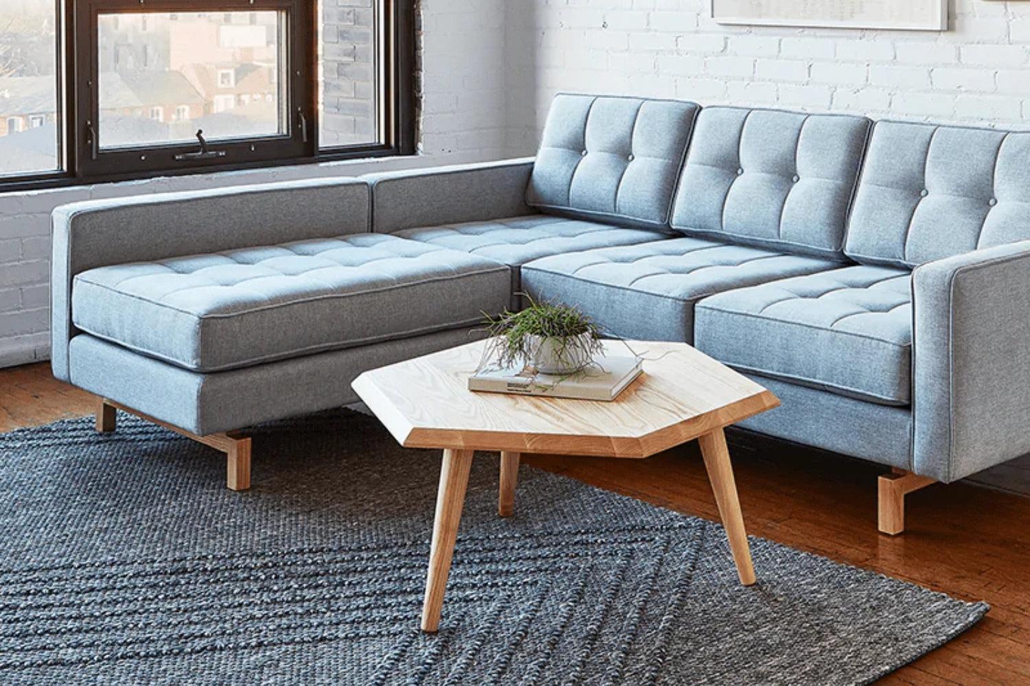 A modern living room features a light gray L-shaped sofa with tufted cushions. In front of the sofa is a wooden hexagonal coffee table with a plant and book on top. The room has a gray textured rug and a large window.