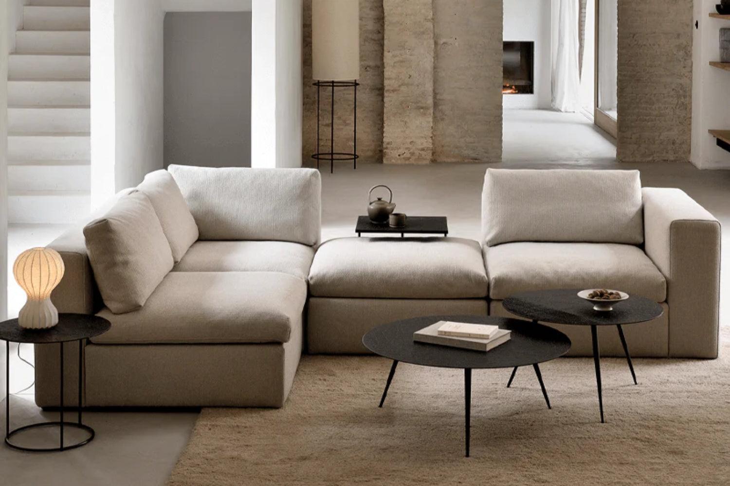 A modern living room featuring a light beige sectional sofa with two black round coffee tables. One table holds a closed book and a small bowl, while the other holds a teapot. A soft, neutral-toned rug covers the floor.