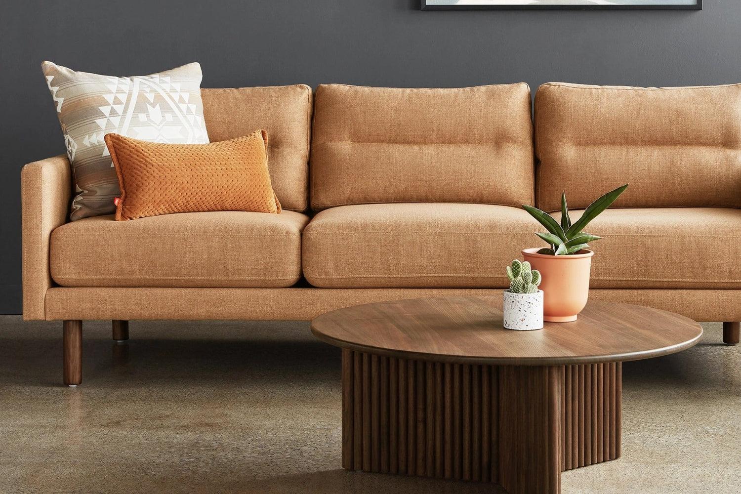 A modern living room with a tan sofa featuring two decorative pillows, one patterned and one textured. A round wooden coffee table in front holds two potted plants. The room has a dark gray wall and a polished concrete floor.