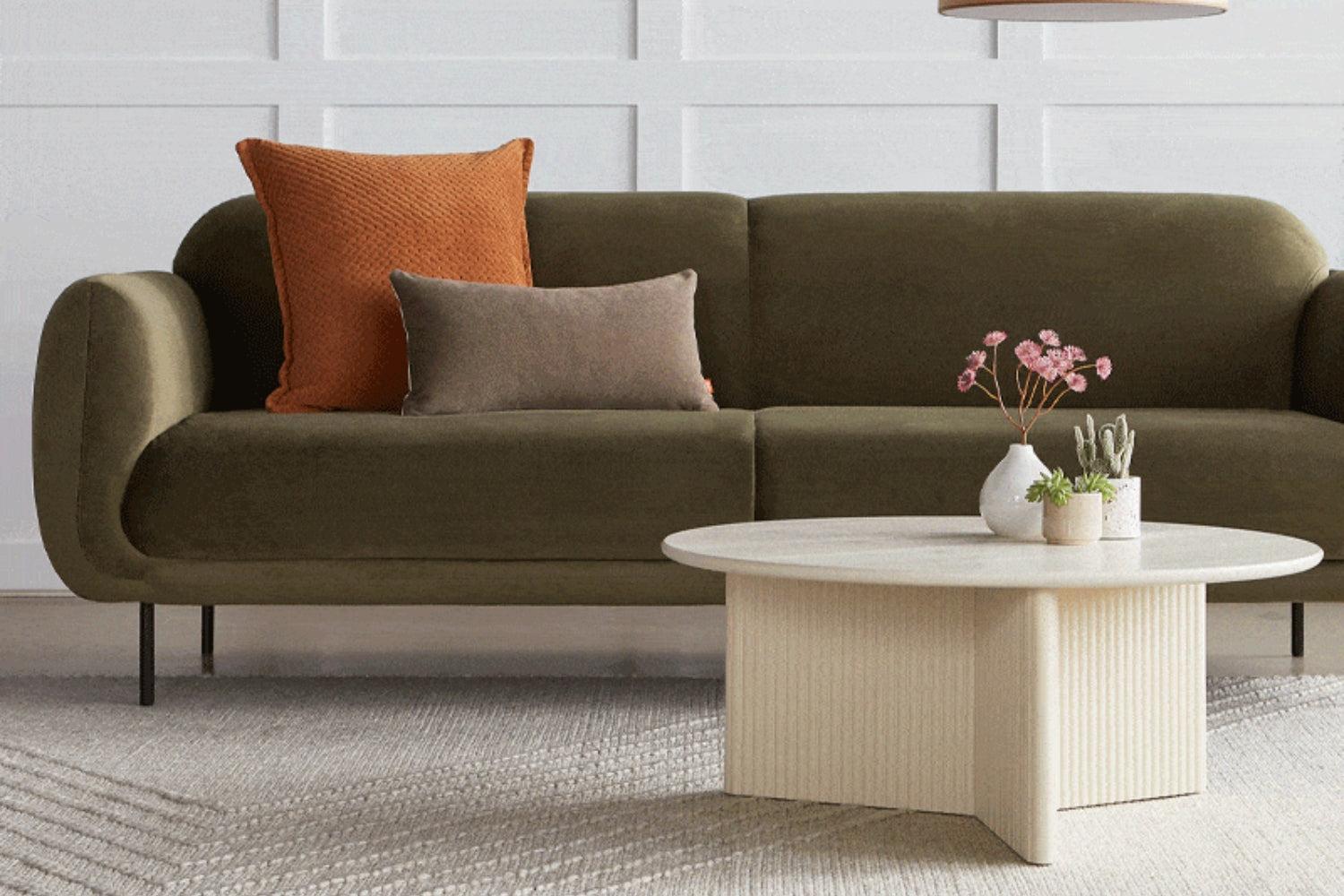 A modern living room with a green sofa adorned with an orange and a gray pillow. A round cream-colored coffee table in front holds small potted plants. The backdrop is a white paneled wall, and the floor is covered with a textured rug.