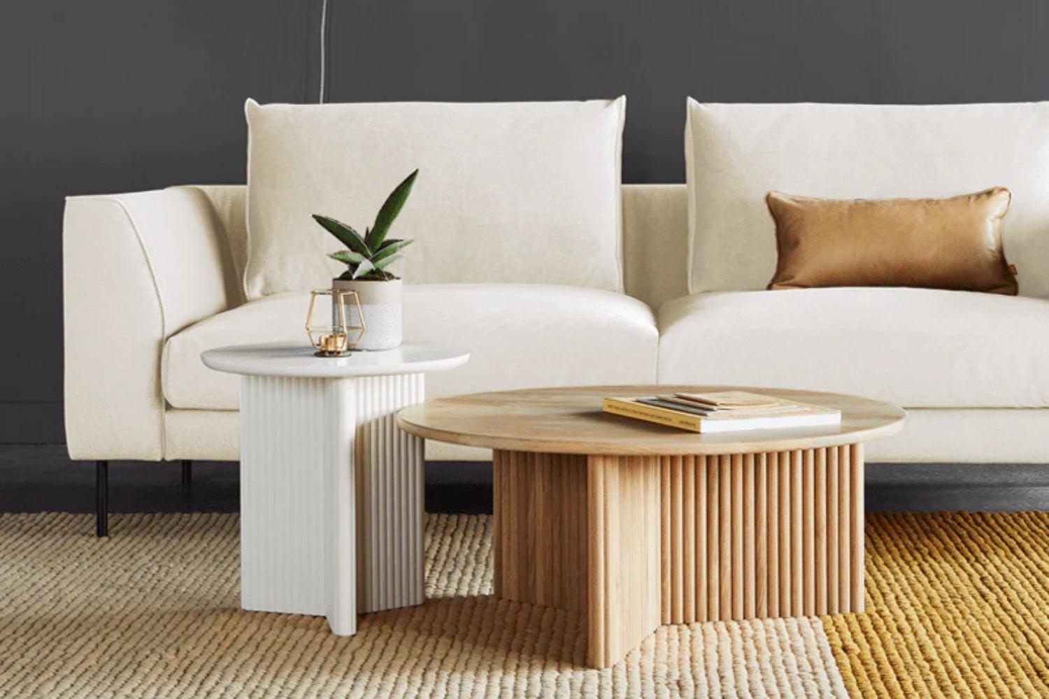 A modern living room with a white sofa featuring a gold pillow. In front of the sofa, there are two round coffee tables; one white with a plant and the other wood-toned with books. The floor is covered with a textured beige rug.