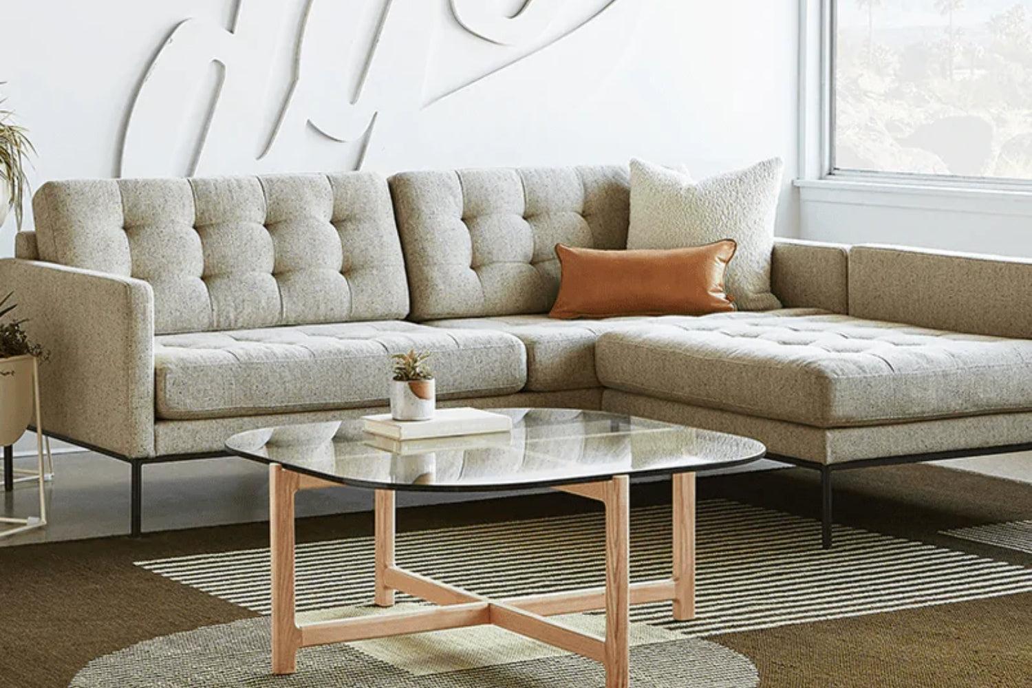 A modern living room with a gray L-shaped sectional sofa featuring tufted cushions. A round glass coffee table with a wooden base sits in front. A white pillow and a brown pillow adorn the sofa. A large window and abstract white wall decor enhance the space.