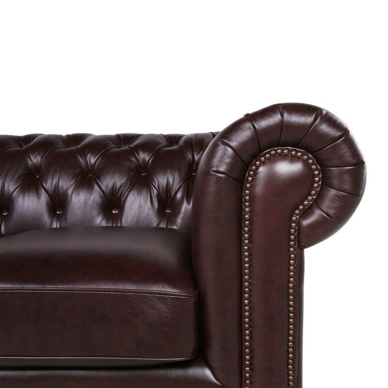 A detailed view of the armrest on the Birch Lane Freddie 95'' Leather Sofa showcases its dark brown, tufted leather with a spiral design and brass nailhead trim. The glossy finish of the luxurious leather accentuates the sofa's elegant, classic style.