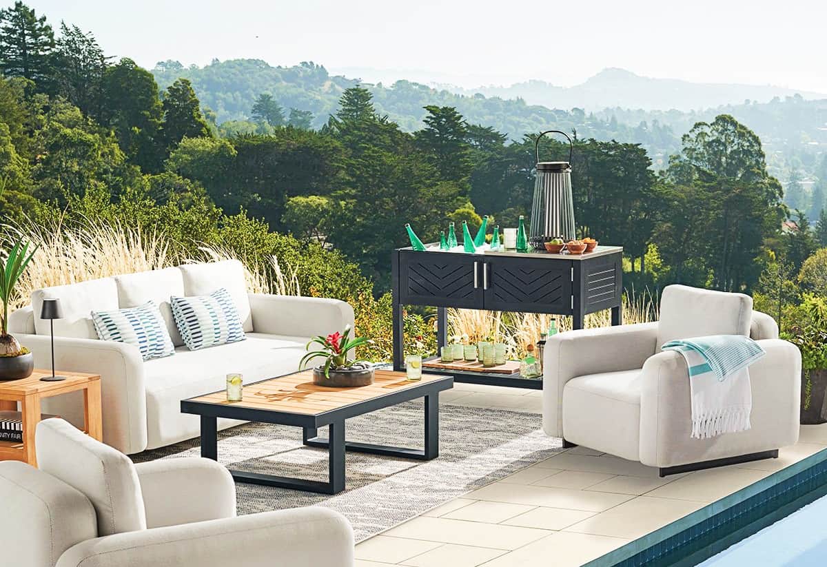 A luxurious outdoor patio setup featuring white cushioned sofas and armchairs arranged around a wooden table with plants and drinks. A mountainous landscape and lush greenery form the scenic backdrop. The area is adjacent to a pool, enhancing the serene ambiance.
