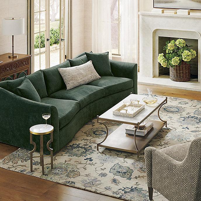 A living room featuring a green curved sofa, a glass coffee table with a gold frame, and a patterned armchair. There's a side table with a drink on it, a rug with floral patterns, and a fireplace with a basket of flowers. Large windows provide natural light.