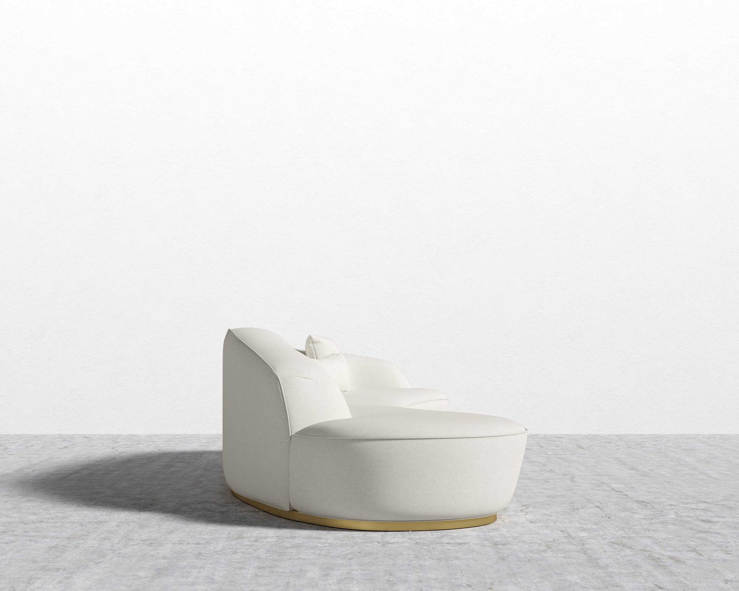 The Reya Curved Sectional, a modern light beige sofa featuring a rounded design and accompanied by a small throw pillow, rests on a gray carpeted floor. It boasts a low backrest and smooth, curved shape, placed against a plain white wall.