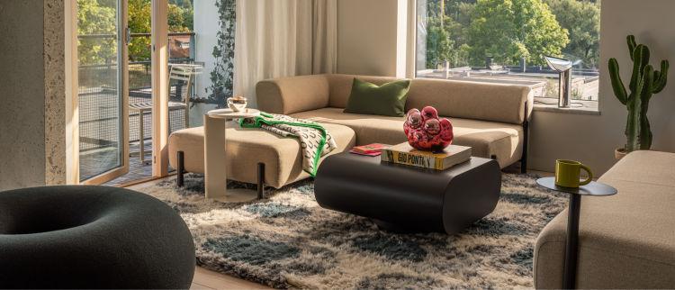 A modern living room with a beige sectional sofa, a black coffee table topped with books and red decorative objects, and a floor-to-ceiling window offering a view of a balcony and greenery. A black round chair, a cactus, and various decor items complement the space.