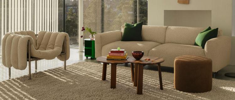 A modern living room with a beige sofa and a unique cushioned armchair, both accented with green pillows. Two wooden nesting coffee tables hold several books and a bowl. An ottoman sits adjacent to the tables, and the room is bathed in natural light from large windows.