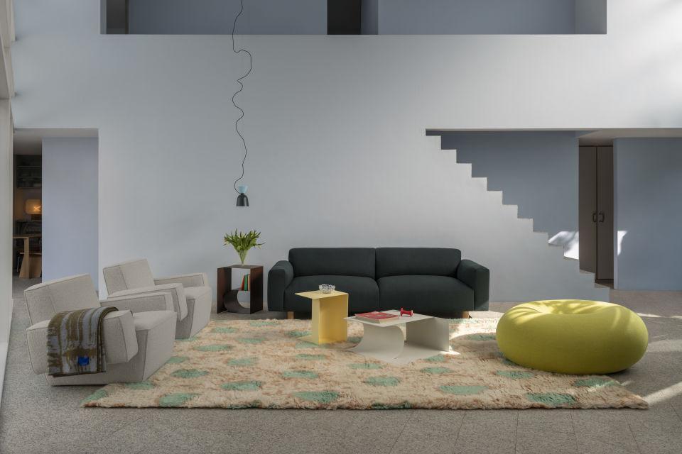 A modern living room features a dark grey sofa, two light grey armchairs, a yellow ottoman, and a geometric rug with pastel patterns. A yellow and white coffee table, a small potted plant, and a pendant lamp adorn the space, with minimalist stairs in the background.