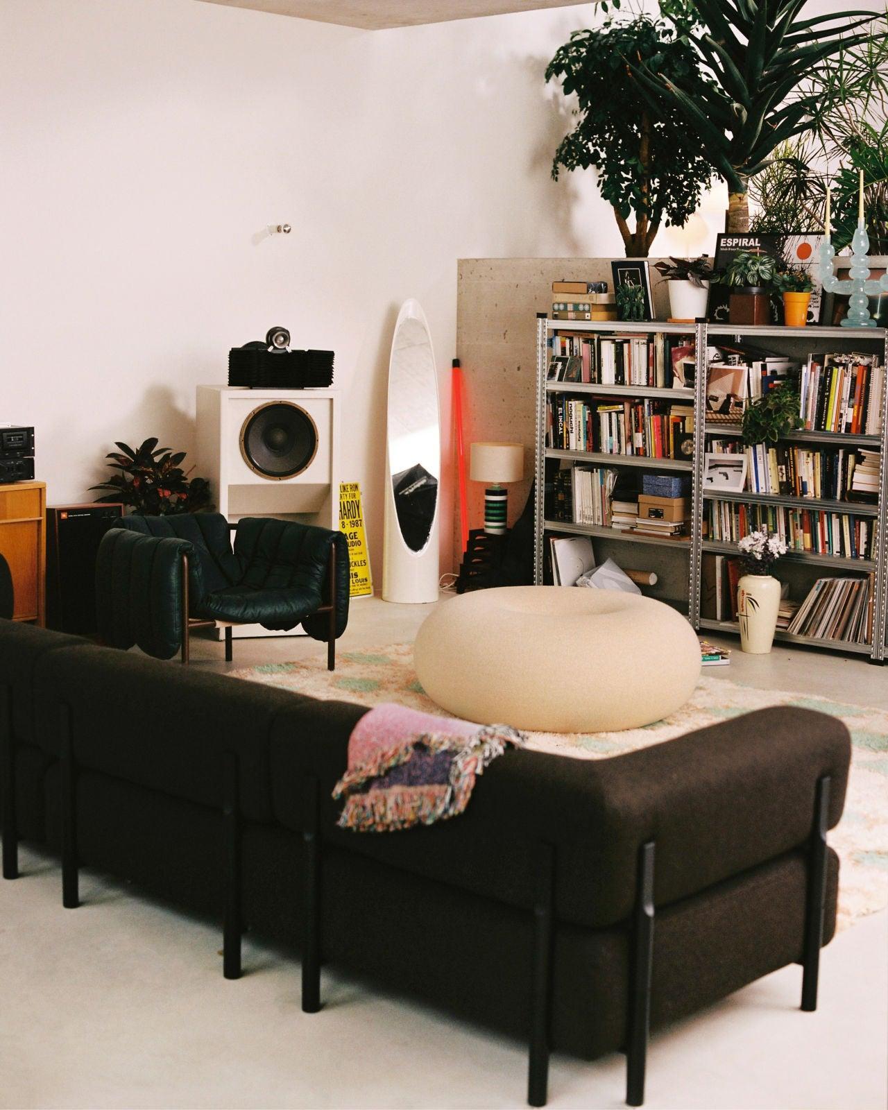 A cozy living room with a dark sofa, a green armchair, a round beige pouf, and a tall mirror. Shelves filled with books and plants, including a tall potted plant, are visible in the background. A speaker and various decor items complete the eclectic setup.
