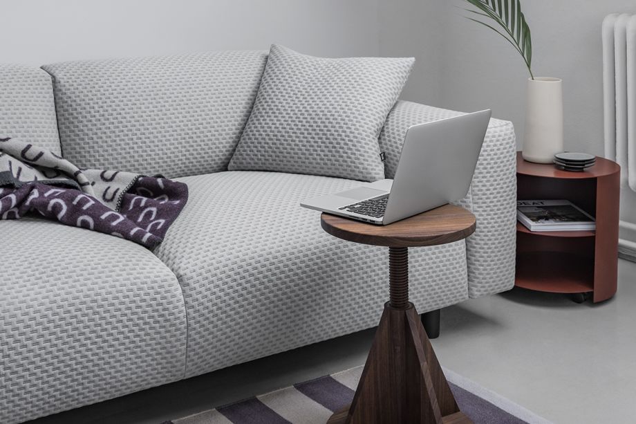 A gray sofa with a textured design has a folded blanket on one end. In front of the sofa is a small, round wooden side table holding an open laptop. Nearby is a red side table holding a white vase with a palm frond and a stack of magazines.