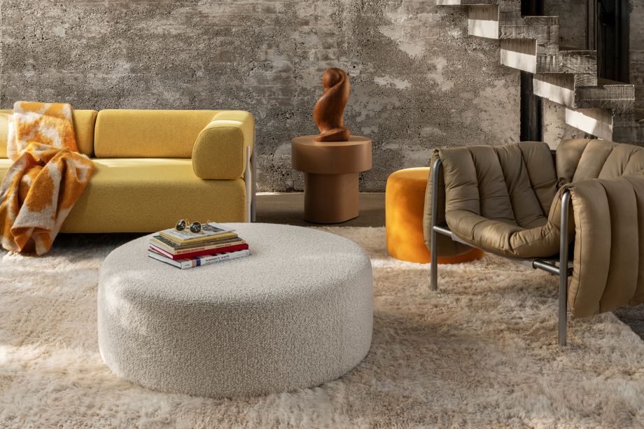 A modern living room with a shaggy beige rug, yellow sofa, brown armchair, and gray circular ottoman with a stack of books on top. A brown abstract sculpture sits on a round table. The backdrop features concrete walls and a staircase.