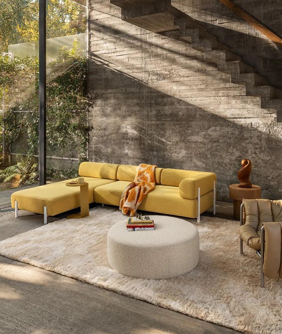 A modern living room featuring a mustard yellow sectional sofa with a white ottoman coffee table in front. A cozy rug covers the floor, and a wooden sculpture sits on a side table. Large windows provide natural light, with a concrete staircase in the background.