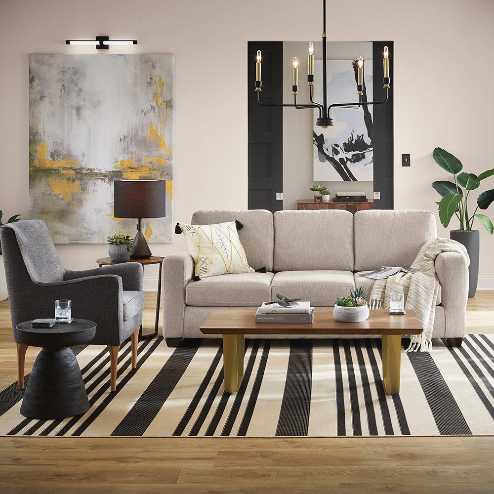 A modern living room features a beige sofa adorned with pillows, a striped rug, a wooden coffee table with decor items, and a gray armchair. The walls display abstract artwork, and a black chandelier hangs from the ceiling. Potted plants add a touch of greenery.