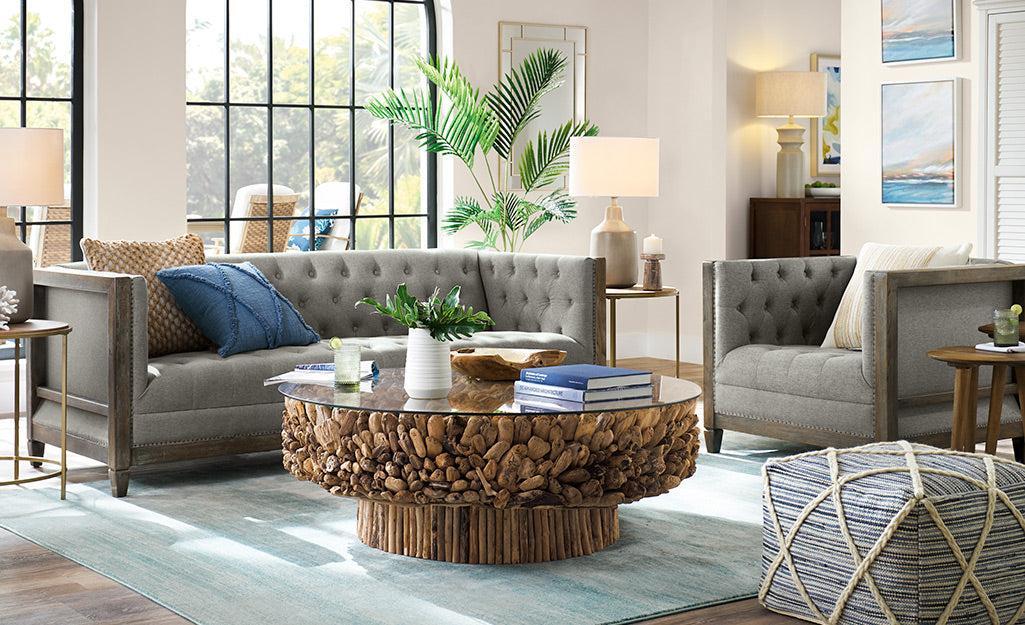 A modern living room featuring a tufted gray sofa and armchair with blue and white accent pillows, a round wooden coffee table with a driftwood texture, indoor plants, table lamps, books, decorative vases, and large windows letting in ample natural light.