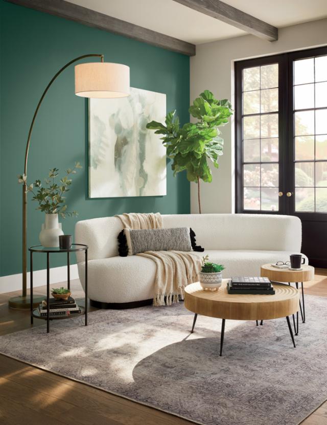 A modern living room with a curved white sofa adorned with a black pillow and a beige throw blanket. A round wooden coffee table and a side table with plants and books sit on a patterned rug. An arched floor lamp and a large plant decoratively complete the space.