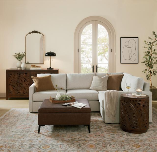 A cozy living room features a white sectional sofa adorned with brown and beige pillows. A brown ottoman serves as a coffee table, and there's a round wooden side table with intricate cut-out designs. The wall holds a minimalist line art piece and tall arched windows.