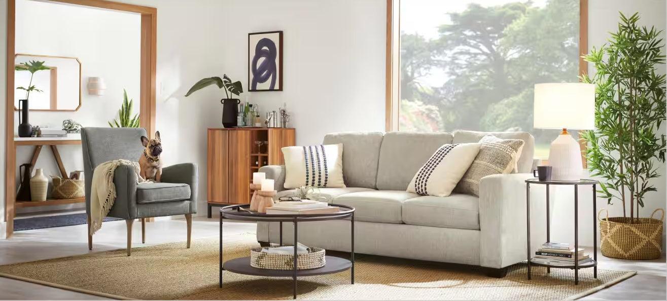 A cozy living room featuring a light-gray sofa adorned with patterned pillows, a round coffee table, and a side table with a lamp. A green armchair with a French Bulldog sits to the left, and large windows reveal a lush, green outdoor view.