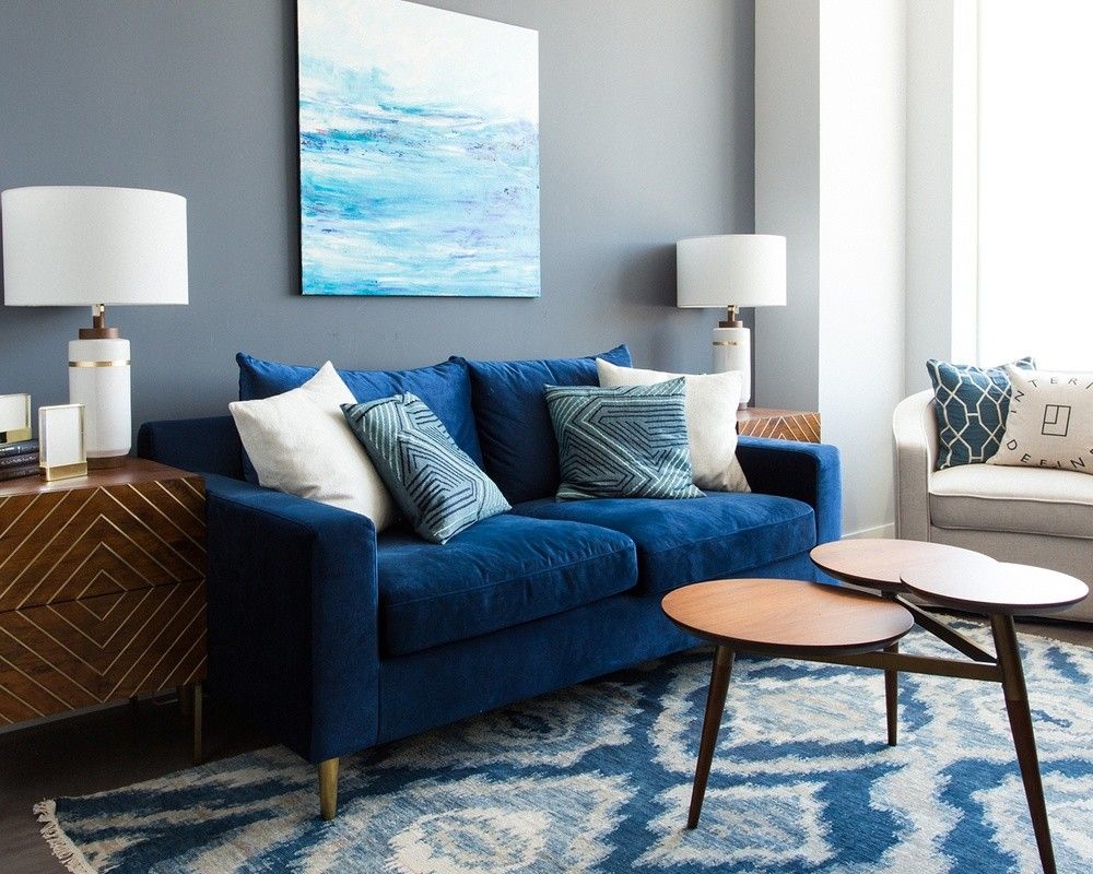 A modern living room with a blue sofa adorned with blue and white cushions. Two wooden side tables with unique designs sit in front of the sofa. A blue and white patterned rug complements the decor. A large abstract painting hangs on the wall behind the sofa.