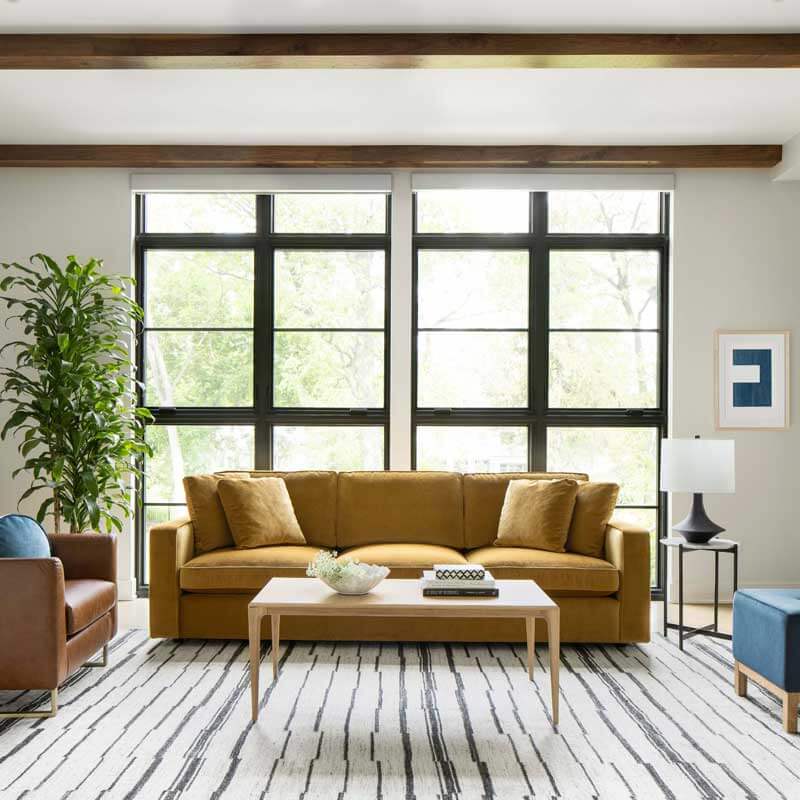 A modern living room features a mustard yellow sofa with matching cushions, a wooden coffee table with a decorative bowl, and a blue armchair. Large windows with black frames let in natural light, illuminating a striped area rug, a tall plant, and framed artwork.