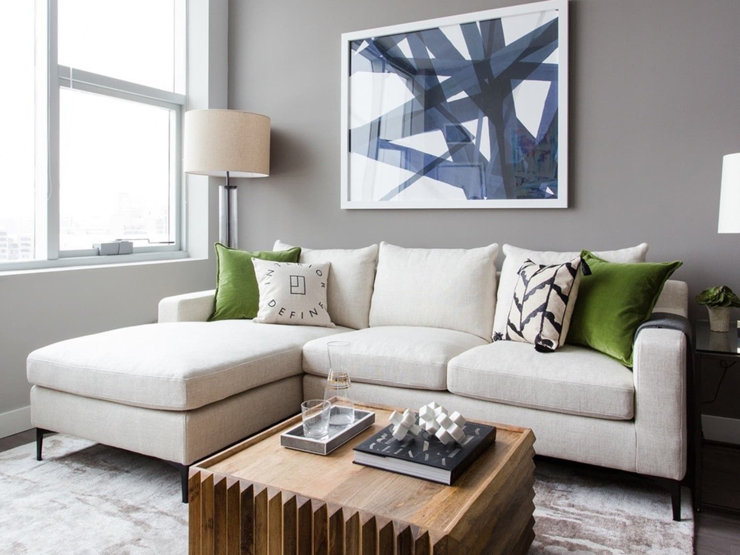 A modern living room with a beige sectional sofa adorned with green and black patterned cushions, a wooden coffee table with decorative items, a tall floor lamp, and a large abstract framed artwork on the gray wall. A window lets in natural light.