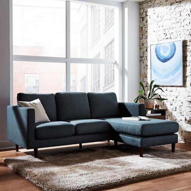 A modern living room with a large window, featuring a blue sectional sofa with a chaise lounge, a beige area rug, and a potted plant on a small wooden table. A blue abstract painting hangs on the white textured brick wall.