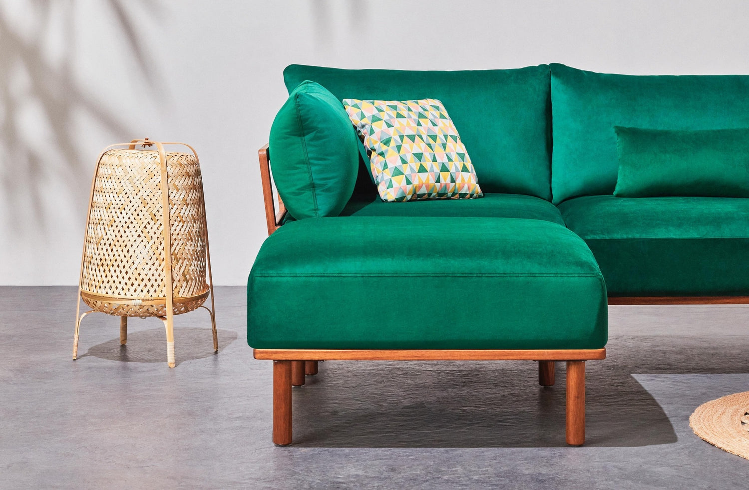 A modern living room setup featuring a green sectional sofa with wooden legs. On the left is a wicker side table topped with a matching lamp. The sofa is adorned with a geometric patterned throw pillow, and the backdrop is a neutral gray wall.