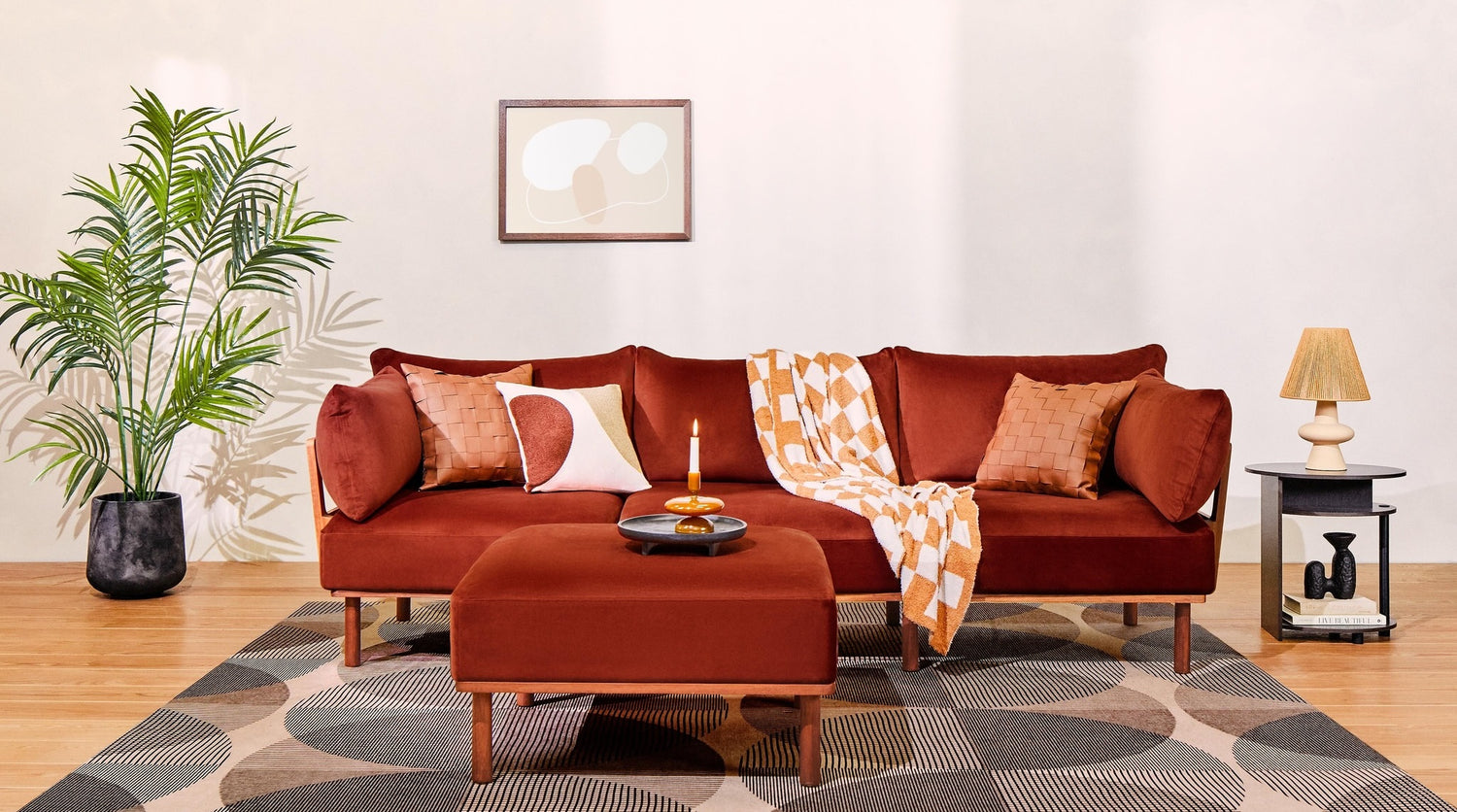 A cozy living room features a burnt orange sofa adorned with patterned pillows and a checkerboard blanket. In front of the sofa, there's a matching ottoman with a candle and tray. A potted plant, framed abstract art, side table with a lamp, and geometric rug complete the scene.