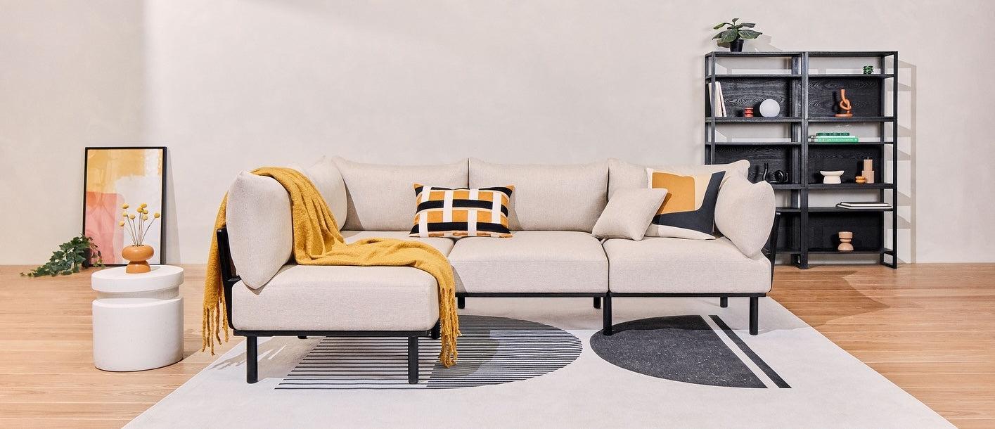 A modern living room features a beige sectional sofa adorned with geometric-patterned cushions and a yellow throw blanket. The room includes a black shelving unit with various decor items, an abstract framed painting, and white cylindrical side tables on a patterned rug.