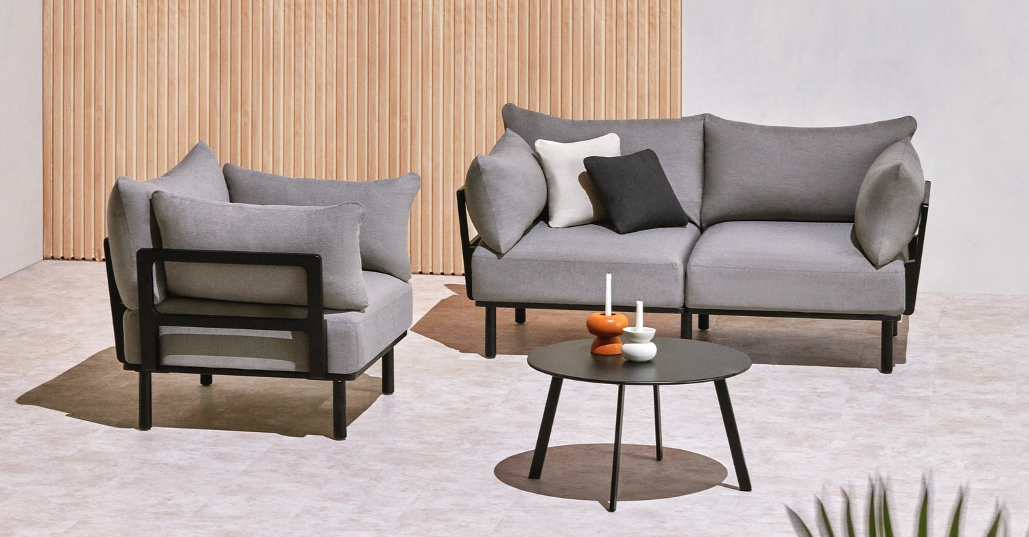 A modern patio setup featuring a grey two-seater sofa with matching armchair, both with black frames. A round, low black coffee table stands in front, holding minimalist decor items including an orange and white object and a small white candlestick.