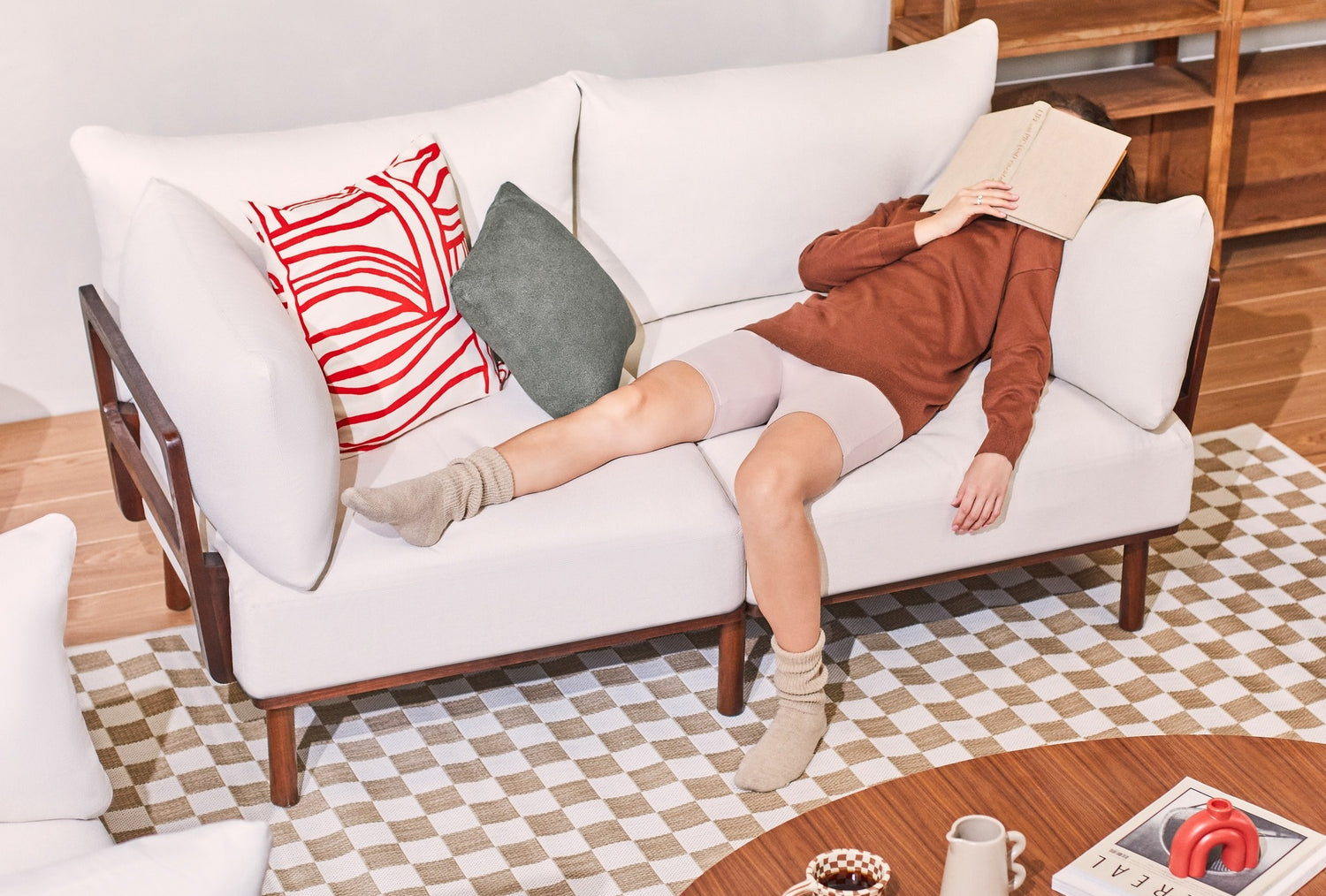 A person is lying on a white sofa with a book covering their face, appearing to be resting or napping. They are wearing a brown long-sleeve top, beige shorts, and beige socks. The sofa has red and green cushions and there is a wooden coffee table in front with various items.