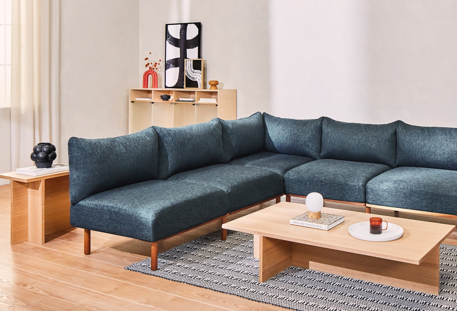 A modern living room features a deep blue sectional sofa with wooden legs. In front is a low wooden coffee table holding a candle, a decorative object, and a book. Beside the sofa is a side table with a sculpture. The background includes a shelf with decor items.