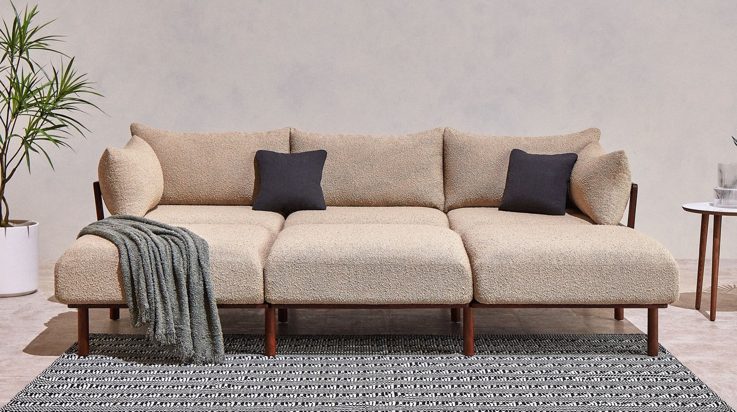 A modern, beige sectional sofa with wooden legs is adorned with two black throw pillows. A gray knitted blanket is draped over the left side. The sofa sits on a black and white patterned rug. A tall plant is on its left, and a small round table with a glass is on its right.