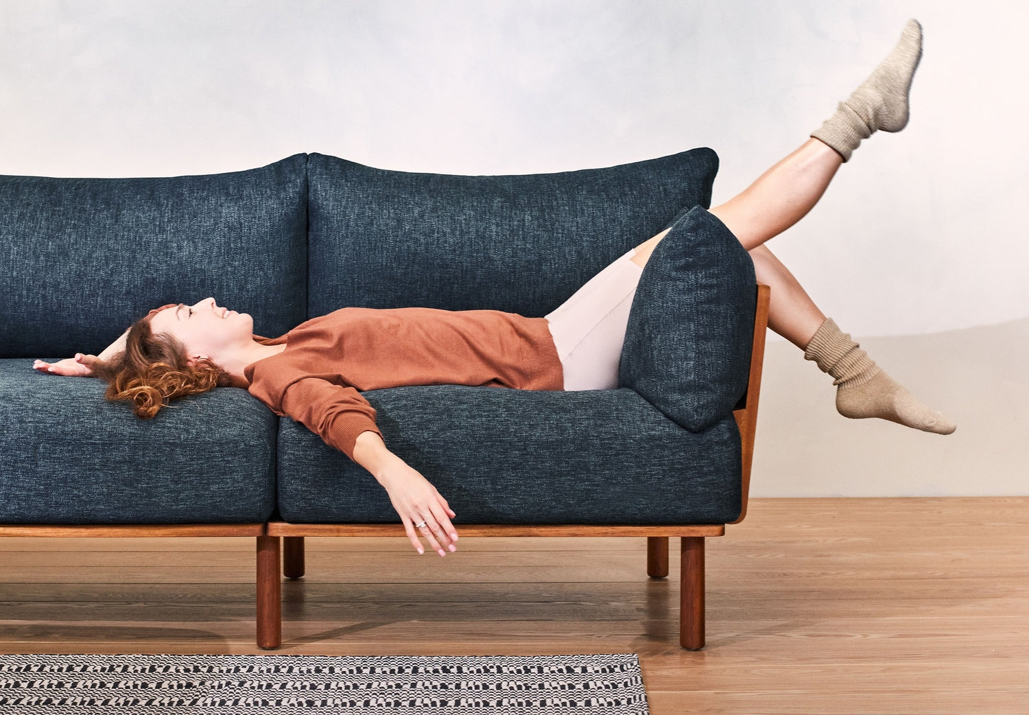 A woman wearing a brown sweater, white pants, and cozy socks is lying upside down on a dark blue sofa. Her legs are resting over the back of the couch, and her arms are stretched out, touching the floor. A patterned rug is on the wooden floor beneath her.