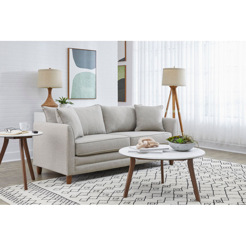 A modern living room features the Kodie 76.5" Upholstered Sleeper Sofa in light gray, accompanied by two floor lamps with wooden bases. Abstract artwork decorates the wall, while a round white coffee table adorned with plants sits atop a patterned rug. Natural light filters through sheer curtains, enhancing the ambiance.