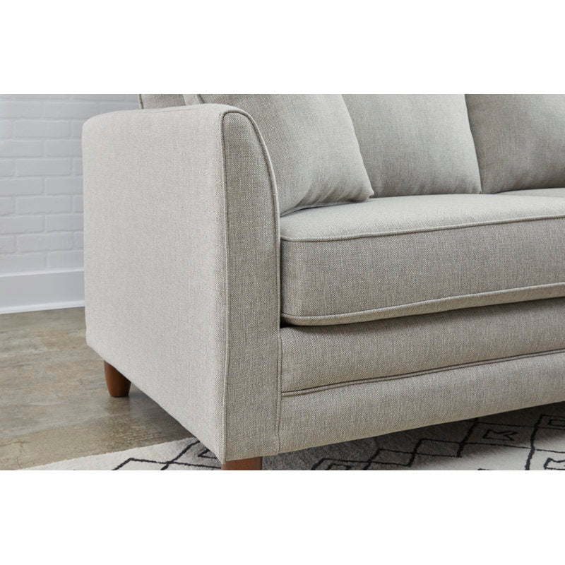 A close-up of the Kodie 76.5" Upholstered Sleeper Sofa in light gray, featuring elegant wooden legs. The sofa is positioned against a white brick wall, with part of a geometric patterned rug visible on the floor.
