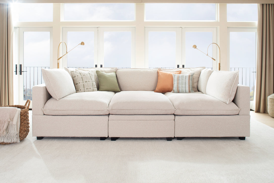 A cozy living room showcasing the Kova Pit, a large beige sectional sofa adorned with patterned and solid-colored throw pillows. Behind the sofa are tall windows with curtains, offering a balcony view, complemented by two modern floor lamps. A woven basket is situated to the left.